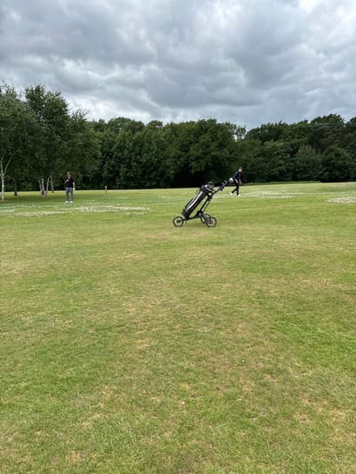 A grassy field stretches into the distance with trees lining the left edge. A golf bag on a wheeled cart is parked on the grass. A person stands near the cart, and another person is walking further down the field. The sky is overcast with heavy clouds.