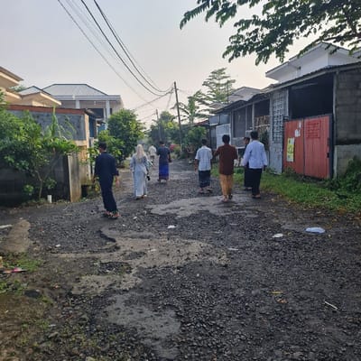 A group of nine individuals, predominantly men, walk along a paved road in Kecamatan Tamansari, Indonesia. The individuals are walking in the same direction, generally moving from right to left in the image.  Most are casually dressed. The individuals are walking on a dark grey, asphalt road with some cracks and a few scattered leaves.  The road is surrounded by greenery, including trees and shrubs. In the background, there are residential buildings, including one with a red door and a sign on the wall.