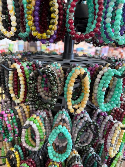 The image displays a close-up, overhead view of numerous beaded bracelets arranged on a multi-tiered display rack. The bracelets are made of small, round beads in a variety of colors including white, black, red, green, yellow, purple, and turquoise. Some bracelets feature a marbled or speckled pattern, while others are a solid color. Silver-colored spacer beads are visible on some of the bracelets. The bracelets are hung on black metal hooks, creating a dense display. The background is blurred, suggesting an indoor environment with other displays of merchandise, possibly a market or shop. There are no people or animals visible in the image. The scene appears to be a retail setting. This specific item is not located in the city of Acıpayam, Turkey.