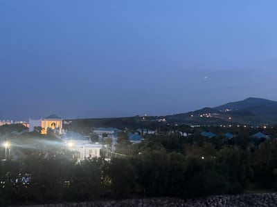 The image presents a serene twilight view of Fnideq, Morocco, capturing a resort or residential area as dusk transitions to night. The sky is a deep, clear blue, with a faint, slender crescent moon visible towards the upper right.
In the foreground, a dark, rocky embankment or wall runs along the bottom, topped by a thick band of trees and bushes, some of which are illuminated by artificial lights, causing noticeable lens flares.
The midground features several buildings characterized by a distinctive architectural style, possibly Moroccan or Andalusian. A prominent, brightly lit building stands out on the left, showcasing intricate arches and design details. Many of the structures have striking teal or turquoise-colored tiled roofs, contrasting with their white or light-colored walls. Scattered artificial lights are visible among the buildings and greenery.
In the background, dark, rolling hills or mountains rise against the horizon. Numerous small, scattered lights dot the slopes of these hills, indicating distant settlements or infrastructure. The scene conveys a peaceful atmosphere as the artificial lights begin to dominate the landscape. There are no visible people or discernible text in the image.