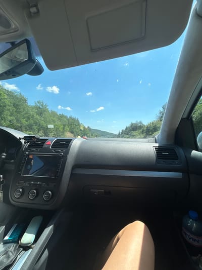 This image captures the interior of a car driving on a sunny day, likely on a road near Rajana, Bulgaria, as indicated by the bottle of "Devin" mineral water visible on the right. The perspective is from the passenger seat, looking forward through the windshield. The dashboard is prominent, featuring a navigation screen, climate controls with red hazard light button, and air vents. A small electronic device is mounted on a holder on the dashboard. Outside, a lush green landscape with trees and rolling hills is visible under a clear blue sky with scattered white clouds. The road ahead appears to be winding. In the foreground, a portion of a person's leg is visible.