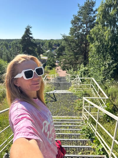 A woman with blonde hair and large white sunglasses stands on a metal staircase in a park setting. She is wearing a pink tie-dye t-shirt. The staircase has white metal railings and is surrounded by green trees and grass. In the background, there are more trees and some buildings visible. The sky is clear and blue. The scene appears to be located in the city of St. Ozerki, Russia.