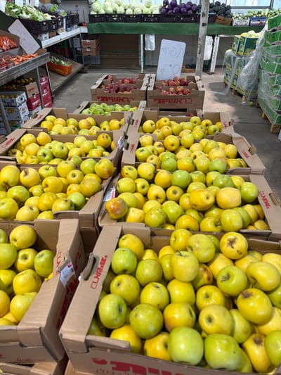 This image captures a market scene in Kadima - Zoran, Israel, showcasing several stacked cardboard boxes filled with yellow apples. The apples are piled high in each box, creating an abundant display. To the left of the apples are other produce offerings, including red cabbages, white cabbages, and other leafy vegetables. There is also a price tag visible near the red apples. In the background, there are more produce items covered in plastic wrap, sitting on pallets. Overall, the scene depicts a typical marketplace setting with a variety of fresh produce available for purchase.