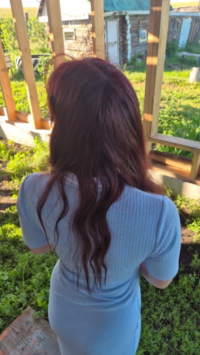 A rear view of a person with long, dark brown hair stands outdoors in Bulatovo, Russia. The person is wearing a light blue, ribbed, short-sleeved dress. They are positioned in front of a wooden structure with vertical beams, beyond which is a grassy area and a rustic log building with a white door and a window. The sun casts shadows from the wooden structure onto the person and the surrounding vegetation.