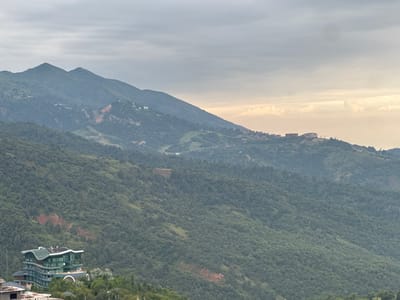 The image captures a scenic view of mountainous terrain covered in dense green foliage, likely a forest. In the foreground, a distinctive modern building with a curved, multi-story structure and a green roof is visible, situated on a hillside. The building appears to be a hotel or resort. Further back, the landscape transitions into rolling hills and then steeper mountains under a cloudy sky. There are scattered signs of human habitation in the distance, with small clusters of buildings visible on the hillsides and mountain slopes. Some infrastructure, possibly ski lifts or roads, can be observed on the mountains. The overall impression is a tranquil, natural setting with a touch of modern development.