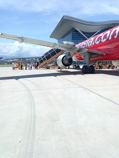 An AirAsia airplane is parked on the tarmac at Cam Ranh International Airport (CXR) in Bắc Cam Ranh, Vietnam. A set of mobile stairs is attached to the aircraft, and passengers are visible boarding or disembarking. The plane's fuselage is bright red with "AirAsia.com" emblazoned in white lettering. In the background, the modern architecture of the airport terminal is visible, featuring large glass windows and a curved roof. The sky is blue with scattered white clouds. The tarmac is a light grey concrete surface with tire tracks. Several ground crew members in high-visibility vests are working around the aircraft. A yellow baggage tractor is also present.