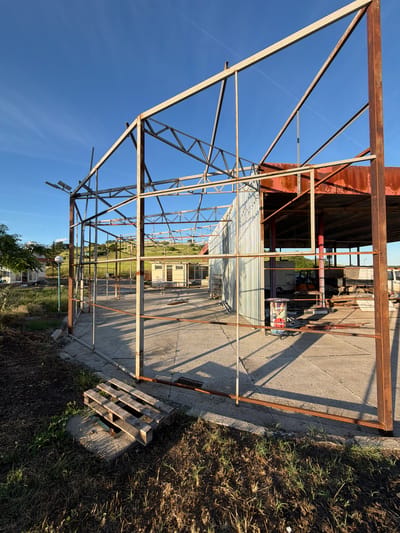 The image depicts a partially constructed metal frame structure, possibly for a greenhouse, located outdoors in Dabnik, Bulgaria. The structure is predominantly composed of rusted metal beams connected to form a series of triangular and rectangular supports. The frame stands on a concrete slab or foundation adjacent to an area with grass and vegetation. In the background, there is a building with corrugated metal siding, as well as other structures and greenery. A wooden pallet rests near one corner of the frame. The sky is visible through the frame, suggesting an open construction. The location is open and appears to be an industrial or semi-industrial area on the outskirts of Dabnik. No people are visible in the image.