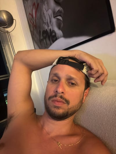 A man with a beard lies down, wearing a black and white eye mask. He has a gold necklace with a cross pendant. His head rests on his left hand. Behind him, there is a wall and a picture frame featuring the word "love". A lamp is visible near the bottom left of the frame. His right arm is exposed, showing his armpit. The scene is lit by natural light, casting a soft shadow across his face and body.