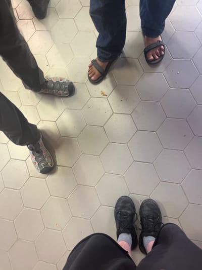 The image presents an overhead, low-angle view of the lower legs and feet of at least four individuals standing on a light-colored hexagonal tiled floor, located within the Brener Regional Council, Israel. The floor tiles are light grey or off-white with visible grout lines, showing minor surface wear and a small, light-brown debris particle near the center.
In the lower foreground, a person is wearing black closed-toe lace-up shoes, light grey socks, and dark-colored trousers or a skirt. To the left, another individual wears dark trousers and grey, orange, and black athletic-style closed-toe shoes with a bungee lacing system. In the upper right portion of the frame, a person is observed wearing dark blue denim jeans and dark grey open-toed sandals, revealing bare feet with visible toenails, some of which appear to have white polish. A fourth individual's dark shoe or boot is partially visible at the extreme upper-left edge of the frame. All subjects appear to be stationary.