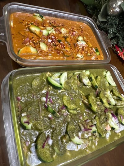 Two glass dishes hold contrasting ceviche variations. The left dish contains a reddish-orange broth speckled with red pepper flakes, diced onion, and sliced cucumber. The right dish presents a green-hued ceviche with sliced cucumbers and diced red onions resting atop a murky green sauce. Shrimp can be seen interspersed throughout the green ceviche. The dishes sit on a wooden surface with holiday greenery nearby.