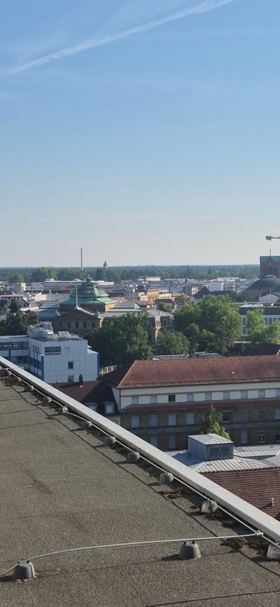 This image captures a view of the city of Karlsruhe, Germany, from a rooftop. In the foreground, a section of a grey, gravel-covered rooftop is visible, equipped with a white railing and several dark, conical anchor points. Beyond the rooftop, the cityscape unfolds. Prominent among the buildings is a large structure with a distinctive green, domed roof, likely a significant landmark in Karlsruhe. Surrounding this central building are numerous other structures of varying architectural styles and heights, interspersed with lush green trees. In the distance, a slender tower or chimney pierces the clear blue sky, which is marked by a faint contrail. The overall scene depicts a sunny day in an urban environment.