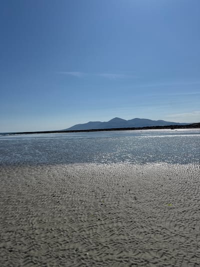 The image shows a wide, sandy beach with gentle waves lapping at the shore under a clear blue sky. In the distance, a range of mountains is visible, silhouetted against the horizon. The sand in the foreground is rippled, indicating the movement of water, and there are small pools of water reflecting the sunlight. A dark, rocky line extends along the coast in the middle ground. The scene is serene and natural, suggesting a peaceful coastal environment. This location is the Ballykinler beach in County Down, Northern Ireland, United Kingdom. There are no people or discernible actions in the image.