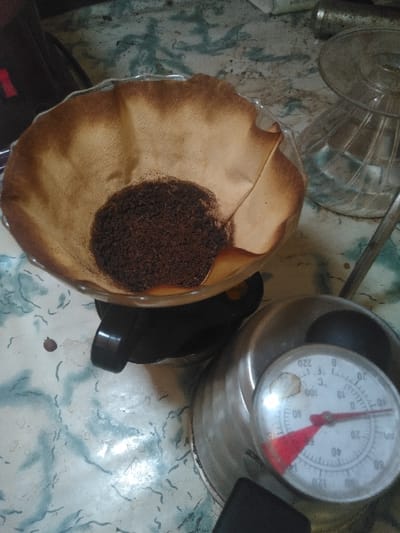 This close-up, high-angle image depicts a coffee brewing setup on a light-colored, mottled countertop in Ciudad de San José, Costa Rica. A conical, light brown paper coffee filter, exhibiting dark brown stains consistent with prior use or moisture, occupies the central left, containing a dark brown mound of coarsely ground coffee. This filter is seated within a clear glass V60-style coffee dripper, which in turn rests on a black plastic base, partially visible beneath. To the lower right, a silver-colored metal gooseneck kettle is partially visible, featuring a black handle. An integrated thermometer on the kettle's lid has a white face with black markings indicating temperatures in both Fahrenheit (0-220°F) and Celsius (0-100°C); its red needle points to approximately 100°C (212°F). In the upper right background, an inverted clear glass dripper or serving carafe is present. The countertop exhibits a wavy, light blue and white pattern with subtle darker specks. No individuals are visible in the scene.