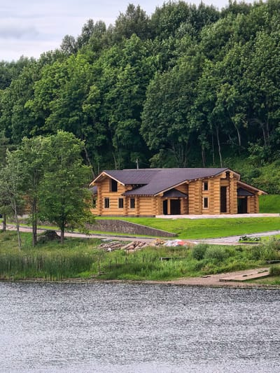 The image depicts a large, modern log cabin situated on a grassy slope overlooking a calm body of water. The cabin is constructed from substantial logs, featuring a dark, pitched roof, multiple windows, and open doorways. The surrounding environment is lush and green, with a dense forest of deciduous trees forming a backdrop to the cabin and a line of reeds growing along the water's edge. In the foreground, the rippling surface of the water reflects the overcast sky. Construction materials, including stacked lumber and what appear to be bags of sand or cement, are visible near the water's edge. The scene is one of a rural or semi-rural setting, likely a residential or recreational property. The location is identified as Staraya Ladoga, Russia. There are no people visible in the image.