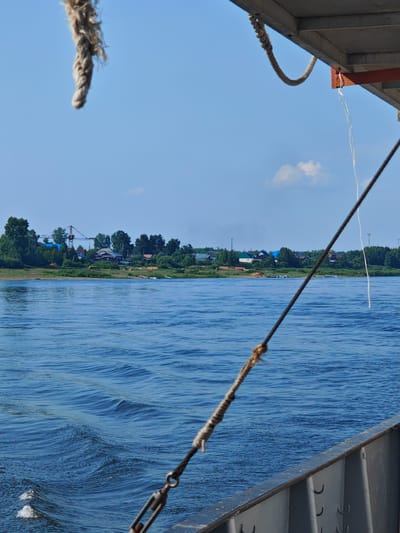 The image depicts a wide, calm river under a clear blue sky with a few wispy clouds. In the foreground, the railing of a ferry is visible, with thick ropes extending upwards and diagonally across the frame. The river's surface is rippled, indicating gentle movement. On the far bank, a tree-lined shore hosts a village, characterized by scattered houses, some with red roofs, and trees. A construction crane is visible behind the village, suggesting ongoing development. The overall scene is tranquil, with the river and sky dominating the composition, and the village providing a human element on the distant bank. The location is identified as Verkhnepashino, Russia.