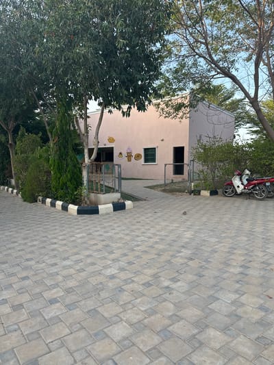 The image depicts the exterior of a small, light pink building with a metal gate and a green-framed window. On the building's facade, there are cartoon drawings of ice cream cones. A pathway made of interlocking pavers leads to the building. To the right of the building, a red and white motorbike is parked. Several trees with green foliage surround the building and the paved area. The scene appears to be set in an outdoor environment, possibly a park or an establishment in Batagarawa, Nigeria, given the context of its location. The lighting suggests it is daytime.