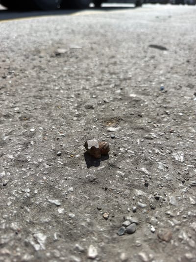 A single, rusty bolt lies on a weathered asphalt surface, casting a distinct shadow to its right. The asphalt is uneven, composed of small gray and white pebbles embedded in darker gray material. The background is blurred, showing what appears to be the edge of a dark tire and a yellow line, suggesting a roadside or parking lot environment. The lighting indicates it is daytime, with bright sunlight. The image is taken from a low angle, close to the ground, emphasizing the texture and detail of the asphalt and the solitary bolt. There are no people or other objects clearly visible in the scene. The location is identified as the city of Lebanon, United States of America.