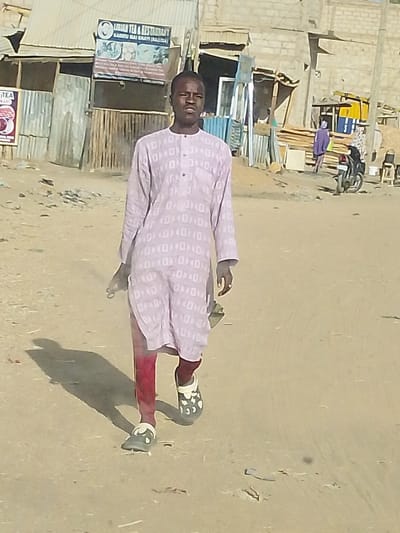 The image depicts a young man walking directly towards the camera on an unpaved, dusty street or path. He is featured prominently in the foreground, taking up a significant portion of the frame. The man is wearing a light purple, long-sleeved tunic (possibly a kaftan or thobe) that extends down to his knees. The tunic has a subtle, repetitive pattern of small, interlocking square-like shapes. Underneath, he wears dark red or maroon pants. On his feet, he has black clogs or sandals with white or light-colored circular and dot patterns. His right arm is slightly bent at the elbow, and his left arm hangs by his side. He has a neutral, somewhat serious expression as he looks towards the camera. A distinct, elongated shadow stretches out behind him and to his right, indicating a strong light source, likely the sun, positioned high in the sky to his left.
The background reveals an urban or semi-urban street scene. To the man's left and behind him are several rudimentary structures, appearing to be small shops or businesses. These buildings are constructed from a combination of corrugated metal sheets (zinc) and light-colored concrete blocks. One building prominently displays a signboard that clearly reads "LIDIAN TEA & RESTAURANT" at the top, with "KADUNA MAI GAYYA (NIGERIA)" written below it. The sign also features graphical representations of food and drinks.
Further into the background, particularly on the right side of the street, more similar structures made of concrete blocks and corrugated metal are visible. Construction materials, including stacks of wooden planks, can be seen near these buildings. To the far right, a person dressed in purple clothing is visible, standing near a parked motorcycle. The ground throughout the scene is sandy or dusty brown, characteristic of unpaved roads in arid or semi-arid environments. The overall impression is that of a bright, sunny day in a busy local area, likely in Nigeria as indicated by the sign.