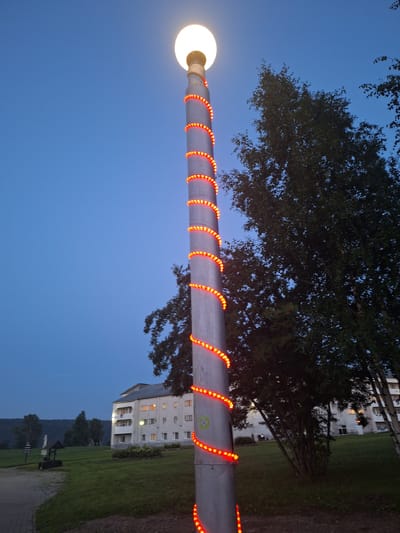The image shows a tall, metal lamppost decorated with a spiral of red LED lights, extending from its base to near the top. A bright white light emanates from the spherical lamp at the very top of the post. The lamppost stands in a grassy area in front of a multi-story building. The building is light-colored with multiple windows, some of which are illuminated. Trees are visible behind and to the right of the lamppost, and a darker treeline is in the background, suggesting a rural or semi-rural setting. A paved pathway leads to the left of the lamppost. The sky is a deep blue, indicating dusk or early evening. The location is identified as Atamanovka, Russia. There are no people or distinct actions depicted in the immediate vicinity of the lamppost.