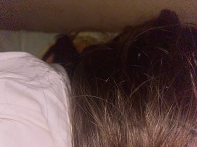 The image is a close-up, overhead shot of what appears to be the back of two people's heads and shoulders, likely in a domestic setting. The foreground is dominated by the dark brown hair of one person, with strands of lighter brown and blonde hair also visible. To the left, a portion of another person's head and shoulder is covered by a white garment, possibly a shirt or bedding. The lighting is dim and appears to be artificial, creating shadows and a grainy texture throughout the image. The background is out of focus but suggests a cluttered or dimly lit environment, possibly a bedroom or a space with objects piled up. There is no discernible information within the image that would indicate a specific location in the city of Afanasyevo, Russia, or any particular activity taking place.