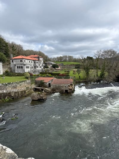The image presents an elevated view of a riverine landscape in A Ponte Maceira, Spain, under a heavily overcast sky. A dark-bodied river flows from the middle right towards the lower left, characterized by turbulent white rapids and a visible weir or dam structure on the far right. Two single-story stone buildings with reddish-orange tiled roofs are positioned on the river's left bank at water level, appearing to be old mills, with one exhibiting partial structural decay near the water. A large, moss-covered boulder rests within the river, positioned centrally. Further up the left bank, a multi-story stone manor house with a prominent reddish-orange tiled roof is situated on a landscaped elevation. This building features numerous windows and is surrounded by manicured lawns, stone retaining walls, and structured hedges. In the background, rolling hills covered with a mix of bare deciduous trees and some evergreen foliage extend under the gray sky. No individuals or active subjects are present in the scene.