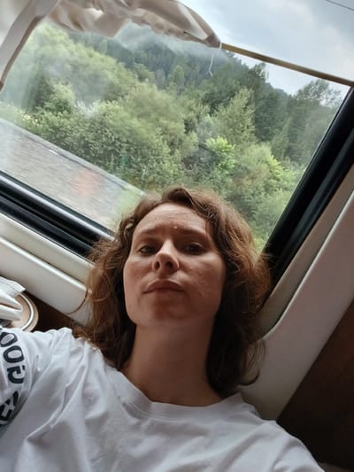 A woman with curly brown hair is taking a selfie inside what appears to be a train carriage. She is wearing a white t-shirt with black lettering on the sleeve. Through the window, a misty, green, forested landscape is visible. The train appears to be moving through the mountains, indicated by the steep, tree-covered slopes and the overcast sky. The interior of the train is visible with white and brown panels. The scene is located in Kolosleika, Russia.