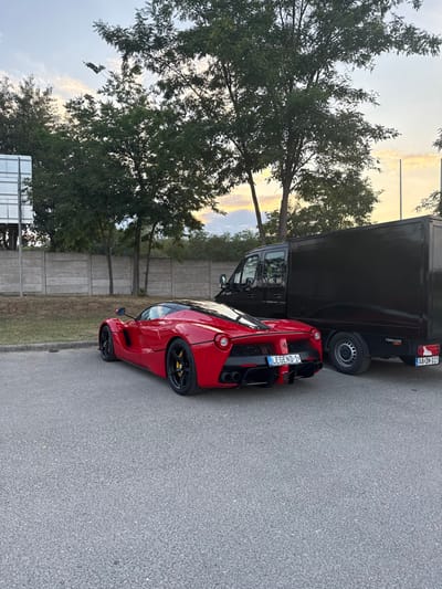 The image captures a static outdoor scene in Mogyoród, Hungary. A red Ferrari LaFerrari, identifiable by its distinct design, is parked on asphalt pavement with its rear facing the viewer and slightly angled towards the left side of the frame. The vehicle features black wheels with yellow brake calipers and displays a personalized Hungarian license plate reading "LEGEND-1." To the right and slightly behind the Ferrari, a black box-bodied panel van, bearing a "TORNADO" sticker and a Hungarian license plate "AA-DN-017," is also parked. In the background, a low, segmented concrete barrier wall stretches horizontally, behind which stands a line of mature deciduous trees with green leaves. The sky above shows a gradient of blue, orange, and pink, indicative of either sunrise or sunset. A partial metal sign structure is visible on the far left, and a single vertical pole is present on the right. No individuals are visible in the scene.