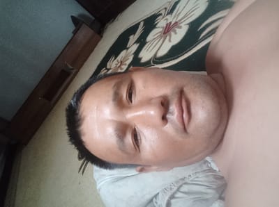 A close-up, top-down selfie of a man lying on his back. He is wearing a light-colored shirt and appears to be indoors. The man's face is the primary focus, with his eyes looking upward. Behind him, a patterned rug with floral designs is visible, along with a wooden cabinet or furniture piece. The overall lighting suggests an indoor environment. The image is a personal photograph, not depicting any specific landmark in Nukus, Uzbekistan.