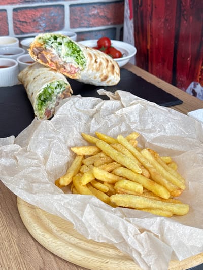 The image shows a wooden platter with two halves of a shawarma-like wrap and a portion of french fries in paper. The wraps appear to contain lettuce, tomato, and some kind of meat. Small white cups containing sauces are also present. A small bowl of cherry tomatoes can be seen in the background. The background shows a faux brick wall and some wood paneling. There is no verifiable information in the image that would indicate a location in Chereshnya, Russia.