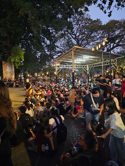 This is a medium shot photograph taken at dusk, showing a large outdoor gathering of people sitting on the ground. The scene appears to be an informal performance or event taking place in a park-like setting in Denpasar, Indonesia. The ground is paved with asphalt, and large, mature trees with dense foliage are visible, casting shadows. A temporary, open-sided structure with a corrugated roof and lighting fixtures is set up in the background, possibly a stage or viewing area. The sky is a deep twilight blue. A tall banner with text is positioned on the left side of the frame. People of various ages are seated, facing towards a central area, some appearing to be watching something. Some individuals are holding phones, suggesting they might be recording or taking pictures. The overall atmosphere is communal and relaxed.