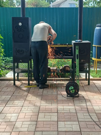 A man wearing a light-colored sleeveless shirt and dark pants is using an angle grinder on a black metal barbecue grill outdoors. Sparks are flying from the grinder, indicating metalworking. The barbecue grill is on a stand with a second, smaller black metal unit next to it. A large blue barrel on a yellow stand is visible in the background. The scene is set on a paved patio with interlocking bricks. Green vegetation and a turquoise corrugated metal fence are also present. The overall environment suggests a backyard or workshop area in a residential setting.