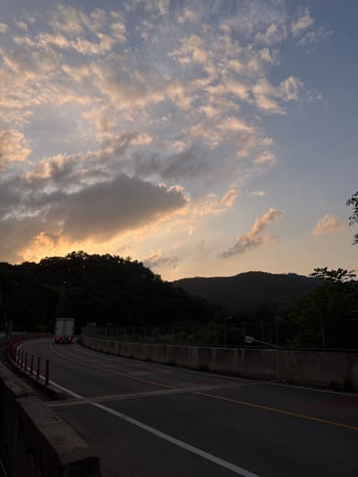 The image depicts a scenic view of a road curving through a mountainous landscape at dusk, likely near Pocheon-si, South Korea. The sky is filled with dramatic clouds illuminated by the setting sun, casting a warm, orange glow on some of them, while others remain in shadow against a blue sky. The road itself is asphalt, with clear yellow and white lane markings. A white truck with its red taillights illuminated is visible in the distance, traveling along the road. The roadside is lined with concrete barriers and orange traffic cones. Dense, dark green trees cover the hillsides, suggesting a natural or semi-natural environment. The overall impression is one of twilight approaching in a rural or suburban area.