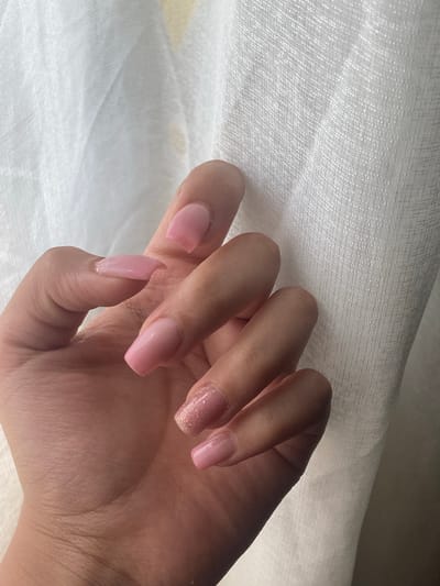 The image displays a close-up of a person's left hand, featuring a fresh manicure. The hand is positioned against a soft, light-colored, textured fabric background, which appears to be a white or off-white sheet or curtain. This fabric shows subtle folds and a fine, woven texture.

The nails are long and shaped in a coffin or tapered square style. Most of the nails are painted with a glossy, solid light pink polish. The ring finger nail stands out with a distinct glittery, sparkly light pink finish, adding a touch of embellishment. The hand's skin tone is light to medium.

The scene is illuminated by soft, diffused natural light, suggesting a daytime indoor setting. The activity depicted is the presentation of the hand, likely to showcase the manicure. There are no overt emotional expressions visible, and no specific location cues within the image directly indicate Topoli, Bulgaria, which is provided as the context. No visible text is present in the image. The overall impression is one of personal grooming and aesthetic detail.