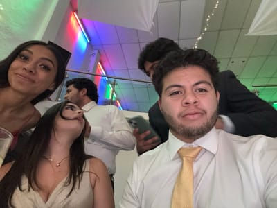 A group of five people are in an indoor setting, likely a party or event. One person takes a selfie, wearing a white shirt and gold tie. To the left, two women are visible, one in a red dress and another with long dark hair wearing a light-colored dress. Behind them, two men are seen, one in a white shirt and the other in a black suit, with one of them holding a smartphone. There is a colored light fixture and decorations in the background.