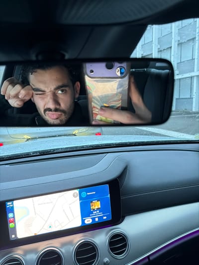 An image captures the interior of a stationary vehicle, looking forward. The primary subject is a male individual reflected in the rearview mirror, taking a selfie. He has dark, curly hair and a beard, winking with his right eye and pouting, with his right fist resting near his temple. His left hand holds a smartphone, encased in a clear or patterned cover, positioned to capture his reflection in the mirror.

The vehicle's dashboard is dark-toned and features a modern infotainment system. The center display shows a split screen: the left panel is a map application zoomed into Mont-de-Marsan, France, clearly labeling "Mont-de-Marsan," "Avenue Pierre Mendès France," and "Parking de la Hourre." The right panel displays a music player showing "Les O" by "Childish Gambino." Below the screen are circular air vents, and a faint purple ambient light strip is visible on the lower right dashboard. Raindrops are present on the windshield, indicating wet weather. Through the windshield and reflected in the mirror, a multi-story building with a light-colored, panelled facade is visible in the background.