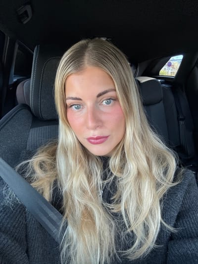 The scene depicts a female individual with long, wavy blonde hair and light-colored eyes, seated in the passenger side of a vehicle located in Meyreuil, France. She is wearing a dark, possibly charcoal grey, knitted sweater with a metallic zipper detail visible at the neckline. A dark grey seatbelt with two prominent horizontal stripes is fastened across her left shoulder and chest. The vehicle's interior features dark grey fabric-covered seats to her left and a black headliner. Behind her, a dark-colored seat is visible. Through the rear side window, a circular road sign with a red border and white center is partially visible, accompanied by blurred green foliage and architectural elements indicative of a street or parking area. The female is positioned looking directly forward, facing the camera.