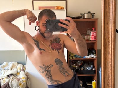 The media file is a selfie taken by a young man in what appears to be a bedroom. The man is shirtless, displaying several tattoos on his chest and torso. He is flexing his bicep and holding up a phone to take the picture, partially obscuring his face. He has dark hair, a beard, and ear tunnels. He is wearing dark blue pajama bottoms.
Behind him, the room is somewhat cluttered. To his left, a bed is visible with white and patterned bedding. To his right, a wooden shelf unit is filled with various items, including bottles, bags, and what looks like kitchenware on top. A tall wooden mirror frame is visible on the far right of the image.
The lighting suggests it is daytime. The overall atmosphere is casual and personal. No specific time of day or weather conditions are discernible. No visible text is clearly legible within the image, apart from a partial label on a red bottle on the shelf that may read "XATA". The setting appears to be domestic.