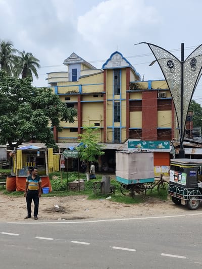 In Benapole, Bangladesh, a three-story building with yellow and red brick facades dominates the background. The building has blue trim around the windows and a decorative, arched top. To the left of the building, a dense green tree stands prominently. In the foreground, a man in a striped shirt and dark pants stands near a roadside stall selling various items. A cart with a covered top, a bicycle, and a rickshaw are also visible in front of the building. The scene is set on a paved road with white lane markings, under an overcast sky.