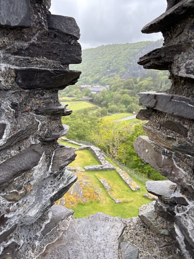 View through a stone embrasure or window of Dolbadarn Castle in Llanberis, United Kingdom. The foreground is dominated by coarse, gray stone construction of the window, with jagged edges and varied textures. Through the opening, a green, grassy area is visible, dotted with stone foundations suggesting historical structures. Beyond the field, a lush, green hillside rises, partially obscured by a misty or rainy atmosphere. A small road or path winds through the foliage, leading to a collection of buildings. The sky is overcast and gray, reinforcing the wet or misty conditions. The perspective is looking down, emphasizing the elevation of the castle.