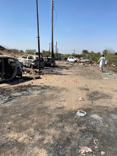 The scene is a street in Tayba Alhasanab, Sudan. The street is covered in dirt and debris, including charred remains of what appears to be a fire. There are several cars parked on the side of the street, some of them burned and charred. A single figure walks away from the scene of the fire in the distance, wearing traditional white clothing. The figure walks away from the camera. There is a utility pole to the left of the image. The environment is dry and dusty, and the sky is blue.