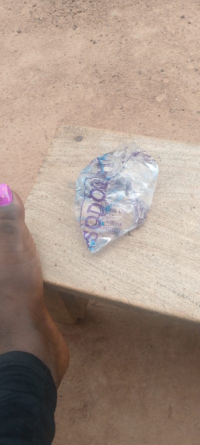 A close-up, eye-level shot shows a plastic bag filled with water resting on a wooden plank. The bag is transparent with purple lettering. The plank is weathered and has a metal tack embedded in it. To the left of the plank, a person's foot is visible, with a pink-painted toenail. The ground surrounding the plank is covered in sandy soil, characteristic of the arid environment found in parts of Adikpo, Nigeria. The lighting suggests it is daytime.
