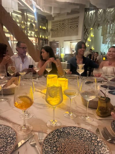 The image displays an indoor restaurant scene with six individuals seated around a large, light-colored marble table. The specific location within El Tezal, Mexico, is not discernible from visual evidence.

From left to right, seated at the table: a person with dark hair, partially visible; an adult male with greying hair and glasses, holding and looking at a mobile phone; an adult female with dark hair, hands clasped on the table, looking to her right; a young adult male with long dark hair, smiling towards his right; an adult female with light-colored hair and a red necklace, smiling towards the camera; and a sixth person, of whom only a hand is visible.

The table is set with multiple stemmed glasses, most containing clear beverages with ice and citrus slices (lemon or orange), and one glass holds an orange-colored drink with an orange slice. Three yellow-glowing cylindrical lanterns are centrally placed on the table, illuminating the immediate area. Ornate, dark-patterned dinner plates are at each place setting, accompanied by silver forks and knives. A basket containing bread rolls is on the left side of the table. A dark, rustic-looking beverage container with a straw and green garnish is on the right.

The background reveals an upscale restaurant interior with warm, ambient lighting. Features include walls with shelves displaying illuminated decorative objects and candles, white fabric draped from the ceiling, a large woven wall hanging, and tree-like decorative structures with small lights. Other blurred patrons are visible in the distant background. The scene suggests a group engaged in dining and social interaction.