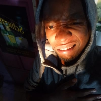 A man wearing a grey hooded sweatshirt is indoors. He is looking at the camera with a slight grimace or strained expression. He has dark skin and a short beard. A sign with "Schwep..." partially visible is in the background. The location is Kinoo ward, Kenya.