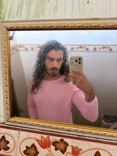 A person with long, dark, curly hair and a mustache is taking a mirror selfie. They are wearing a pink long-sleeved shirt. The mirror is framed with ornate gold and cream detailing. The background appears to be a tiled bathroom. The person is holding a light-colored phone with a camera bump, which is reflecting in the mirror. The walls in the reflection are tiled with a pattern of pink and brown floral motifs. The overall environment suggests an indoor setting, likely a bathroom, in Selemadeg Barat, Indonesia.