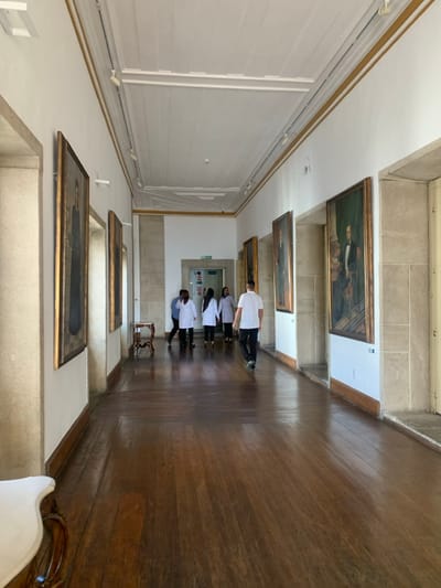 The image displays a long, well-lit interior hallway within the Palácio da Aclamação, located in the Campo Grande neighborhood of Salvador, Bahia, Brazil. The hallway features polished dark wooden floorboards, white painted walls, and a high ceiling with white wooden paneling and ornate molding accented with gold-colored trim, equipped with track lighting fixtures.
Multiple large, framed oil portraits are mounted along both walls. On the left wall, two portraits are visible, the foremost depicting a dark-robed figure. A small, dark wooden console table with ornate legs stands against the left wall further down the corridor. On the right wall, at least three large portraits, depicting men in formal attire, are displayed within recessed, light-colored stone frames. Similar stone frames define doorways or architectural recesses along the corridor.
Seven individuals are present. In the mid-ground, a group of six walks away from the camera, deeper into the hallway. Five members of this group wear white lab coats, while the individual leading them wears a light blue shirt and dark pants. Separately, a male individual wearing a white short-sleeved t-shirt and dark pants walks towards the camera on the right side of the hallway, distinct from the group. At the far end of the hallway, a doorway is visible with a green "EXIT" sign positioned above it. In the immediate foreground, bottom left, a portion of a white-upholstered, ornately carved wooden furniture piece is visible.