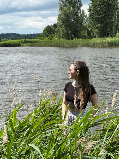 In the city of Вішоўскі сельскі Савет, Belarus, a young woman with long brown hair and glasses stands near the water's edge, partially obscured by tall green reeds and dry stalks of vegetation. She is wearing a black crop top and light-colored shorts. Her hair is pulled back with a gold hair clip. She is looking to her right, away from the camera, with her left hand resting on her hip. The background shows a large body of water with ripples, and beyond that, a treeline composed of various green trees under a cloudy sky. The lighting suggests it is daytime, likely overcast or a partly cloudy day.