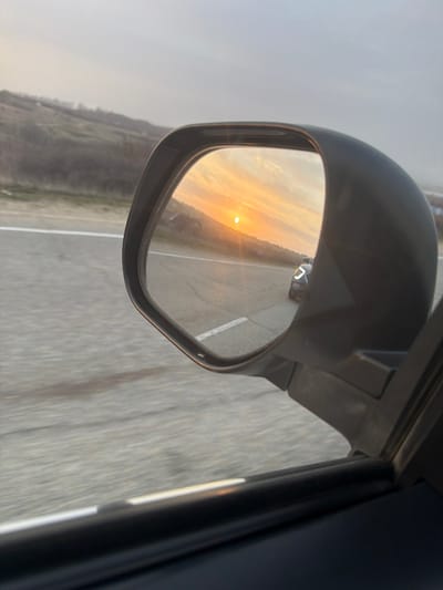 The image presents a view from inside a moving vehicle, near Gostagaevskaya, Russia, focused on its passenger-side exterior mirror. The black-framed mirror prominently reflects a sunset or sunrise, with the sun, a distinct orange-yellow disc, low on the horizon. The sky in the reflection exhibits streaky clouds bathed in warm golden and orange light. A dark-colored vehicle, possibly a sedan or SUV, with illuminated headlights or daytime running lights, is visible behind the observer's vehicle in the mirror's reflection, traveling on the same asphalt road. Outside the mirror, the gray asphalt road with a faded white edge line shows motion blur, indicating the vehicle is in transit. Rolling, sparsely vegetated hills are visible in the distance under a hazy, overcast sky. The dark frame of the car's window and interior trim are visible in the foreground.