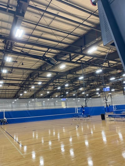 The image shows the interior of a large gymnasium with a wooden floor. The gymnasium is equipped for volleyball, with nets and poles visible. Blue padding is around the poles, and blue curtains hang behind the nets. The ceiling is high and industrial, with exposed beams and ductwork. Numerous fluorescent lights illuminate the space. In the foreground, the polished wooden floor reflects the overhead lights. Along the side of the court, there are bleachers and a trash can. A scoreboard with the number "7" is visible in the distance. The scene appears to be unoccupied by athletes or spectators. The overall impression is of a well-maintained sports facility.