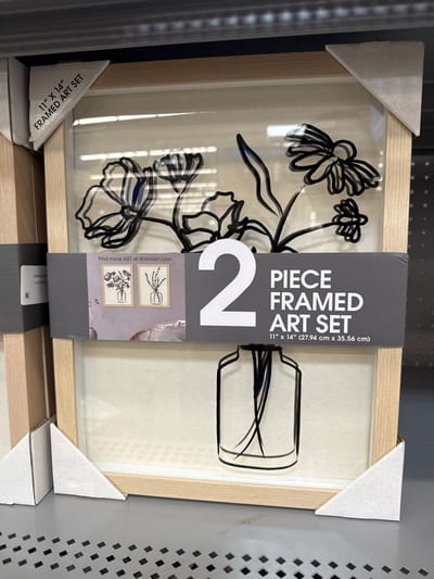 A retail product shot displays a "2 PIECE FRAMED ART SET" on a store shelf, viewed from a slightly upward angle. The art set is rotated about 90 degrees clockwise. Each piece features minimalist black line art on a light, off-white textured background, encased in a light wooden frame.

The left art piece depicts a bouquet of abstract flowers with multiple petals and leaves. The right piece shows a simple vase containing a few slender, curved lines resembling reeds or minimalist branches. Both pieces are protected by a clear covering, which reflects ambient light.

The two framed artworks are held together by a dark gray cardboard band, which serves as packaging. White cardboard corner protectors are visible on the frame corners.

Visible text on the packaging includes: "2 PIECE FRAMED ART SET", "11" x 14" (27.94 cm x 35.56 cm)", and "Find more ART at Walmart.com". Small thumbnail images on the label suggest other available art pieces or variations. The setting appears to be a retail environment, with a black perforated metal shelf visible on the right side of the image.