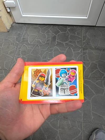 The image shows a hand holding a stack of colorful cards with Lego characters on them. The cards have a red and yellow border, and on them are images of Lego figures, one a warrior with a sword and the other two figures in futuristic suits. The scene appears to be outdoors, with a tiled ground and a white door or gate in the background. The location is identified as Suceava, Romania.
